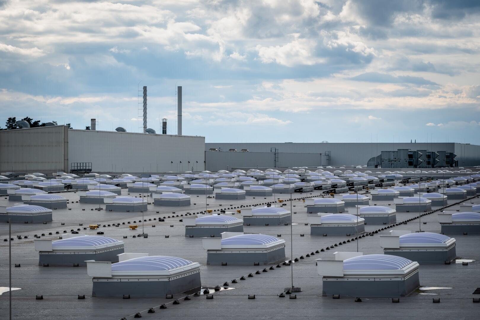 Ein großes Industriedach mit zahlreichen kuppelförmigen Oberlichtern unter einem bewölkten Himmel, mit einem Fabrikgebäude und hohen Schornsteinen im Hintergrund.