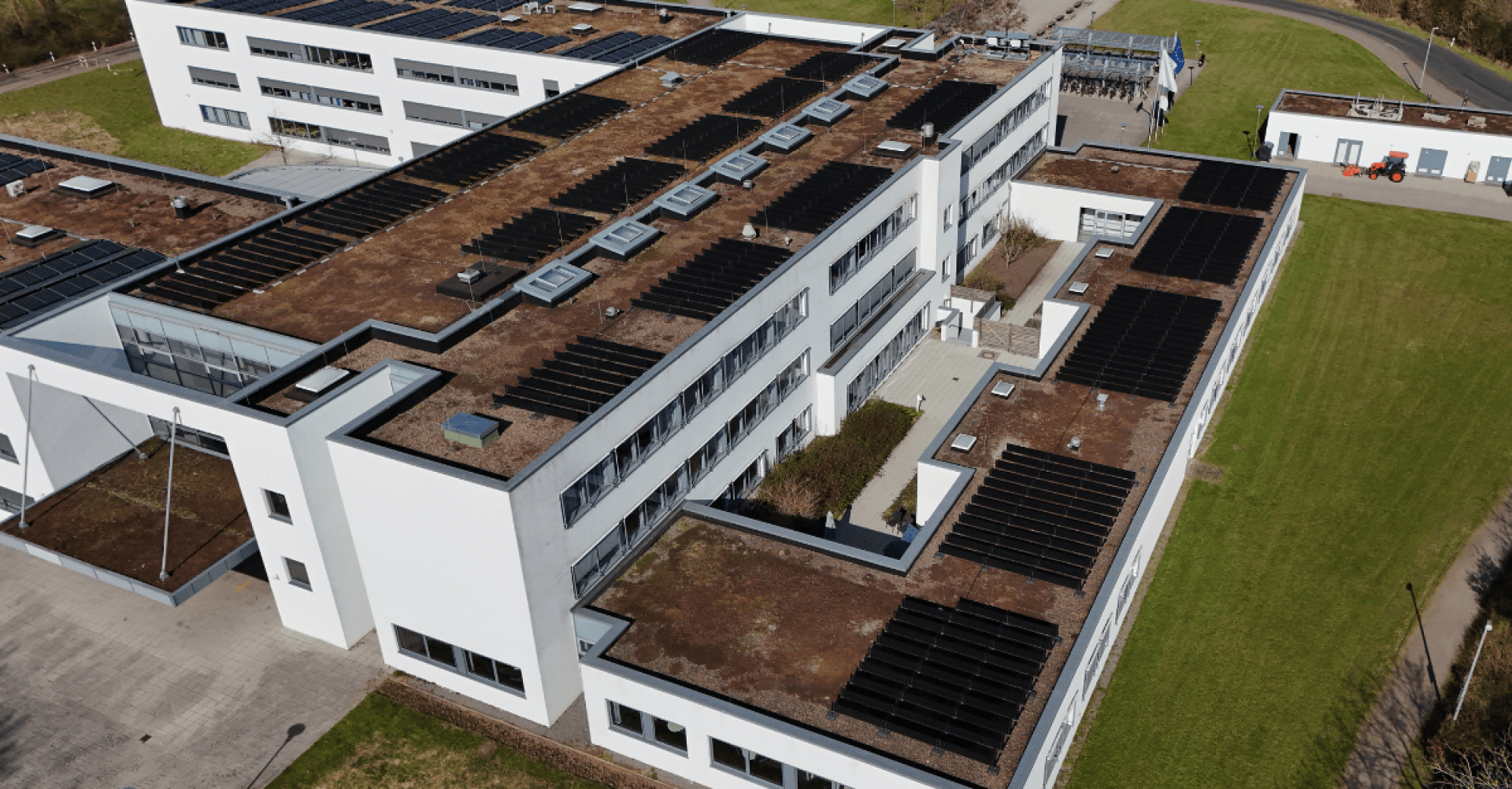 Aerial view of a modern white building with several floors, a flat roof covered with solar panels and skylights, surrounded by lawns and paved paths.