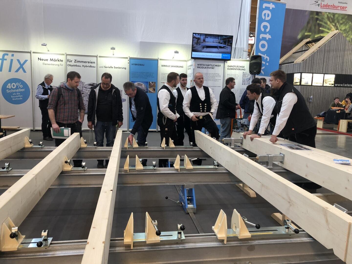 Eine Gruppe von Personen, einige in traditioneller Zimmermannskleidung, steht um große Holzbalken und Metallmontagegeräte auf einer Messe oder einem Ausstellungsstand. Im Hintergrund sind Informationsbanner und ein Bildschirm zu sehen.