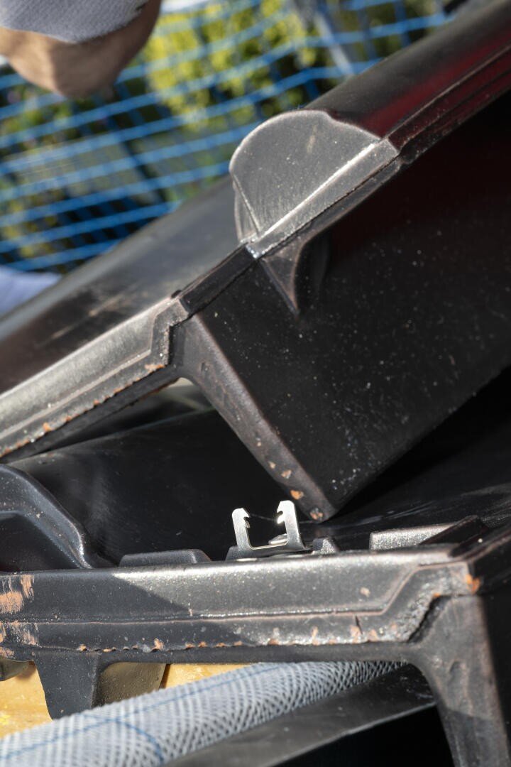 Nahaufnahme von gestapelten schwarzen Dachziegeln mit einer Metallklammer dazwischen, oben ist ein Teil einer Hand zu sehen, und im Hintergrund ist ein blaues Sicherheitsnetz zu erkennen.