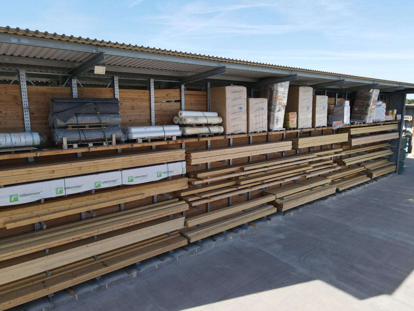 Stacks of wooden planks and building materials are organized on metal shelves under a corrugated metal roof in an outdoor storage area, with boxes and rolls stored on the top shelf.