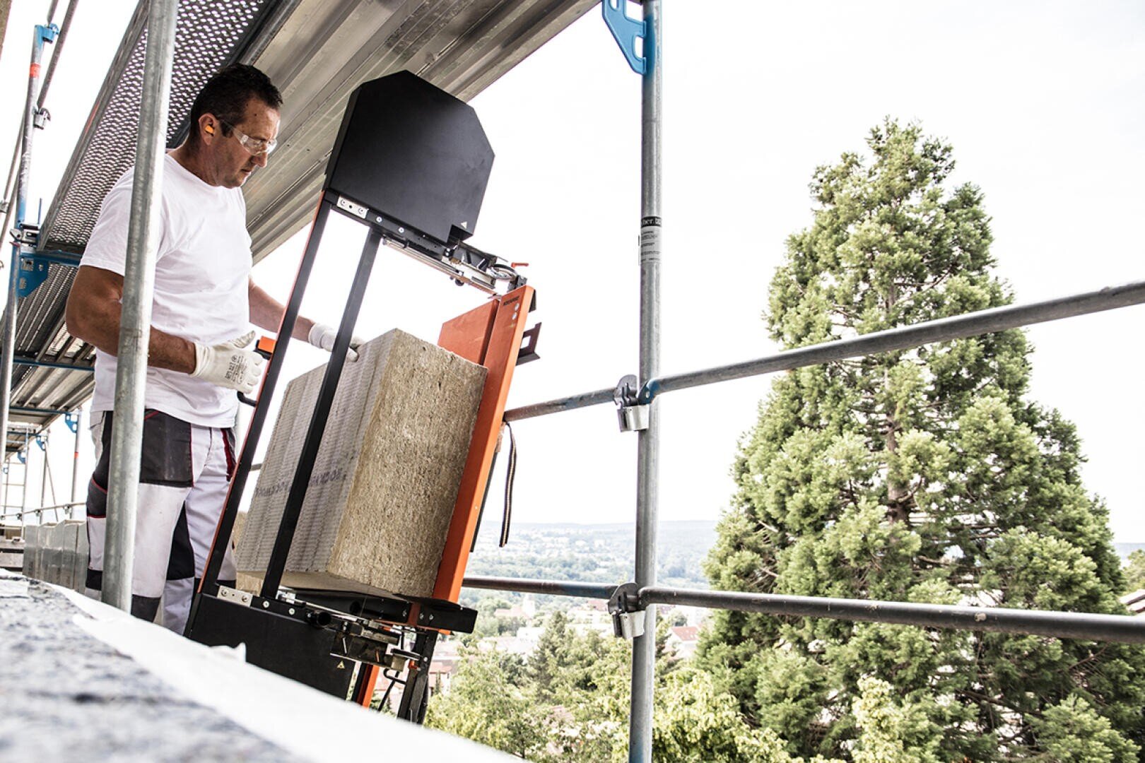 A construction worker in white clothing operates a machine to cut stone blocks on scaffolding. A large tree and a distant landscape can be seen in the background.