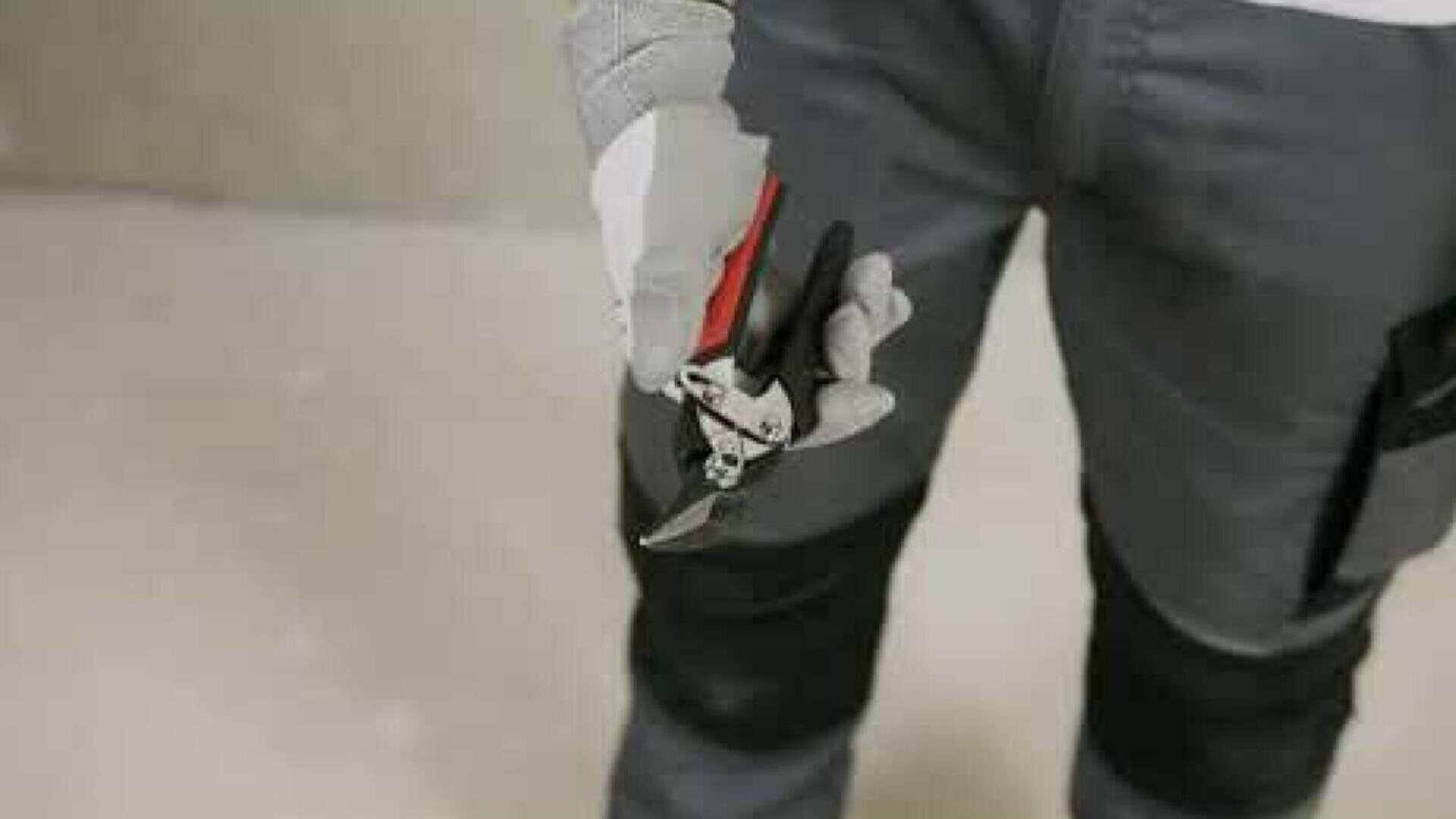 A person wearing gray pants and work gloves holds a pair of metal pruning shears with a red handle, standing indoors on a light-colored floor.