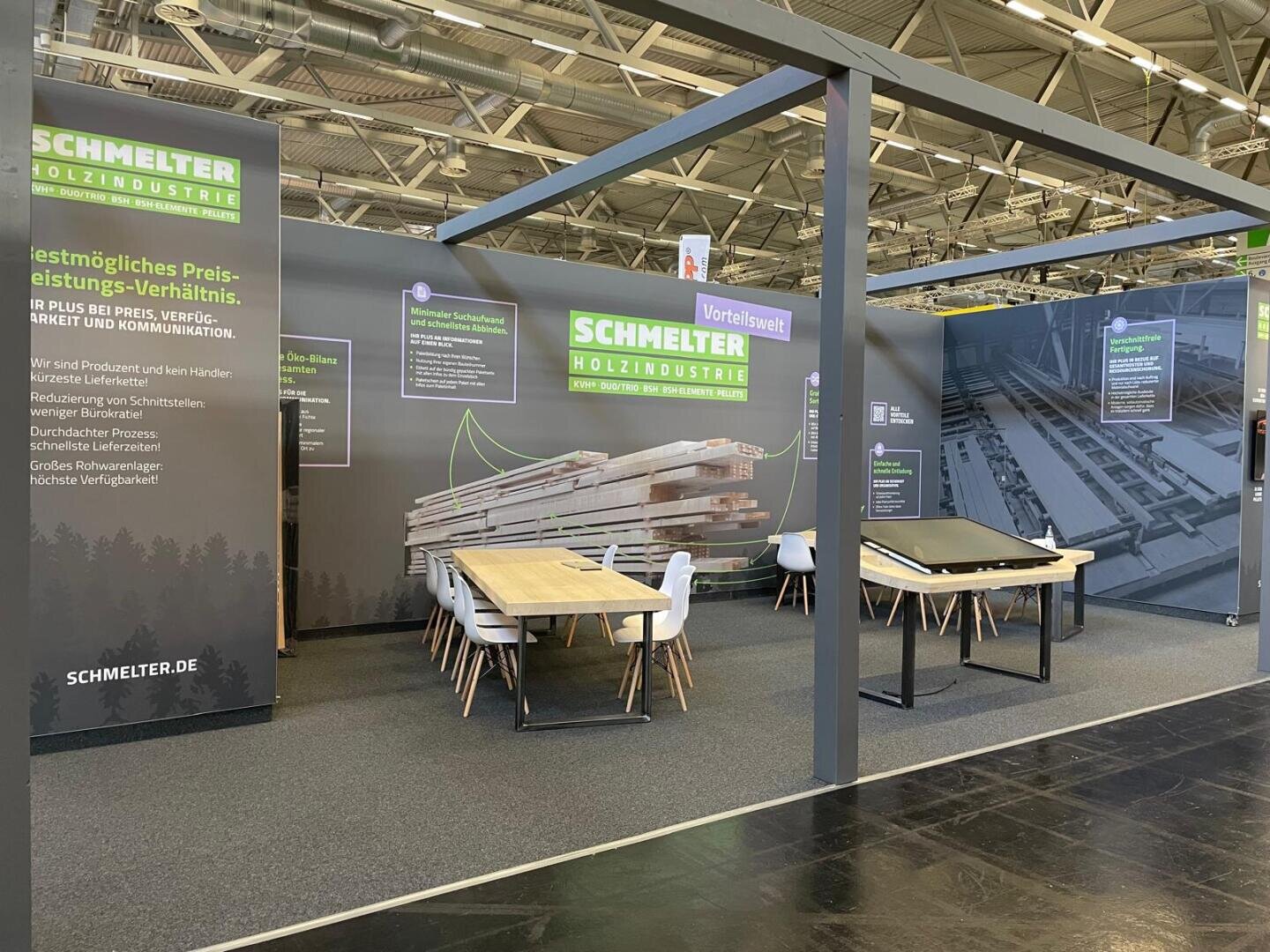 Messestand für Schmelter Holzindustrie mit großen Bannern, Tischen, Stühlen und gestapelten Holzgrafiken. Der Stand ist in einer geräumigen Halle mit Oberlichtern und Informationsdisplays aufgebaut.