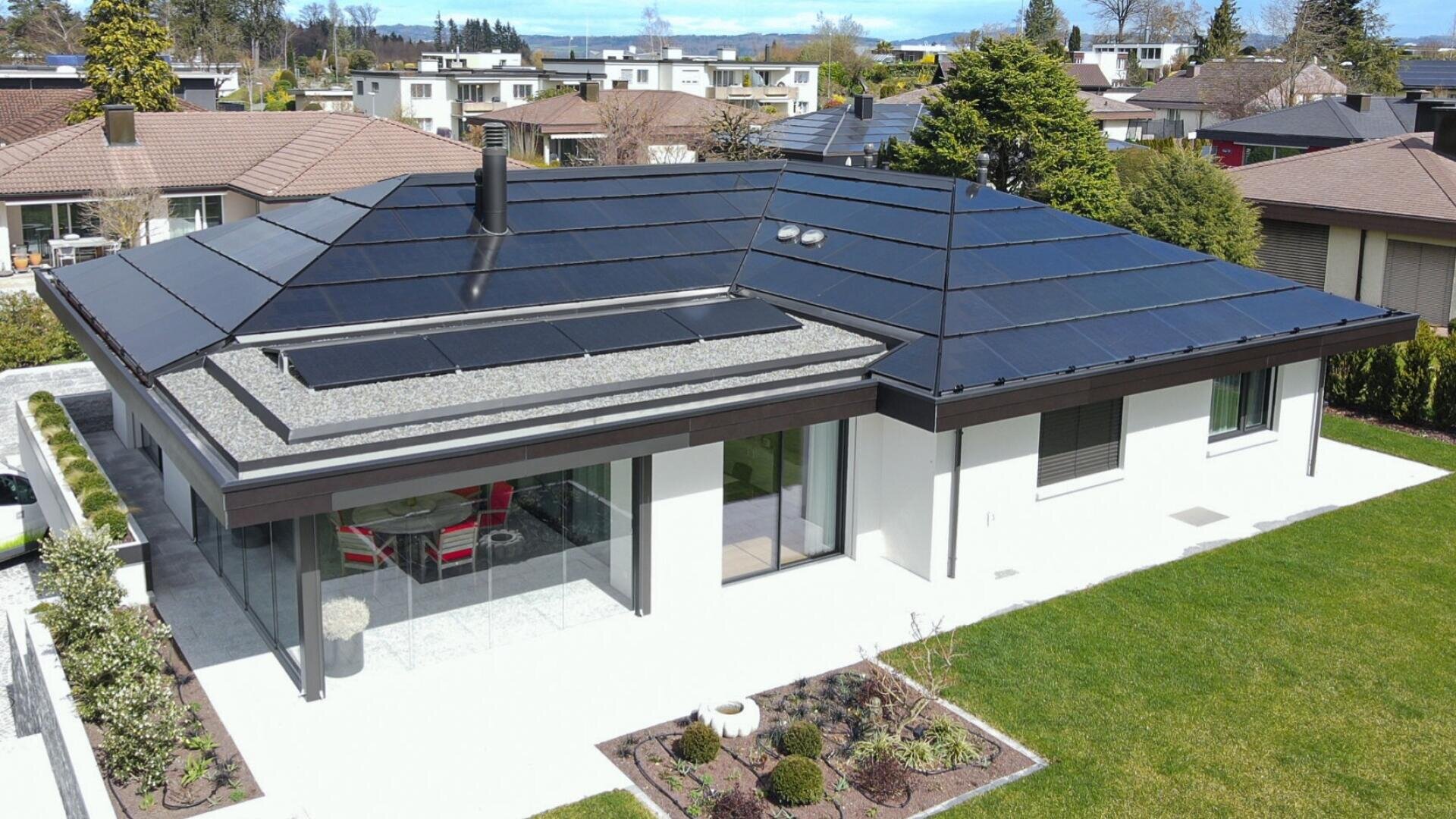 Modern single-story house with large windows and black solar panels integrated into the roof, surrounded by a well-maintained lawn, small garden, and neighboring homes under a partly cloudy sky.