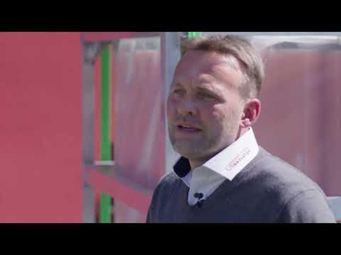 Ein Mann mit kurzen Haaren, einem weißen Hemd mit Kragen und einem dunklen Pullover steht im Freien vor einem modernen Gebäude mit roten und gläsernen Scheiben.