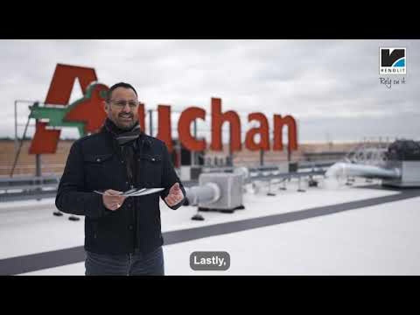 Ein Mann steht auf einem Dach und hält ein Klemmbrett in der Hand, hinter ihm ein großes rotes Auchan-Schild. Der Himmel ist bewölkt, und man sieht Industrieanlagen. Der Untertitel lautet: "Lastly", und in der rechten oberen Ecke erscheint ein Logo.