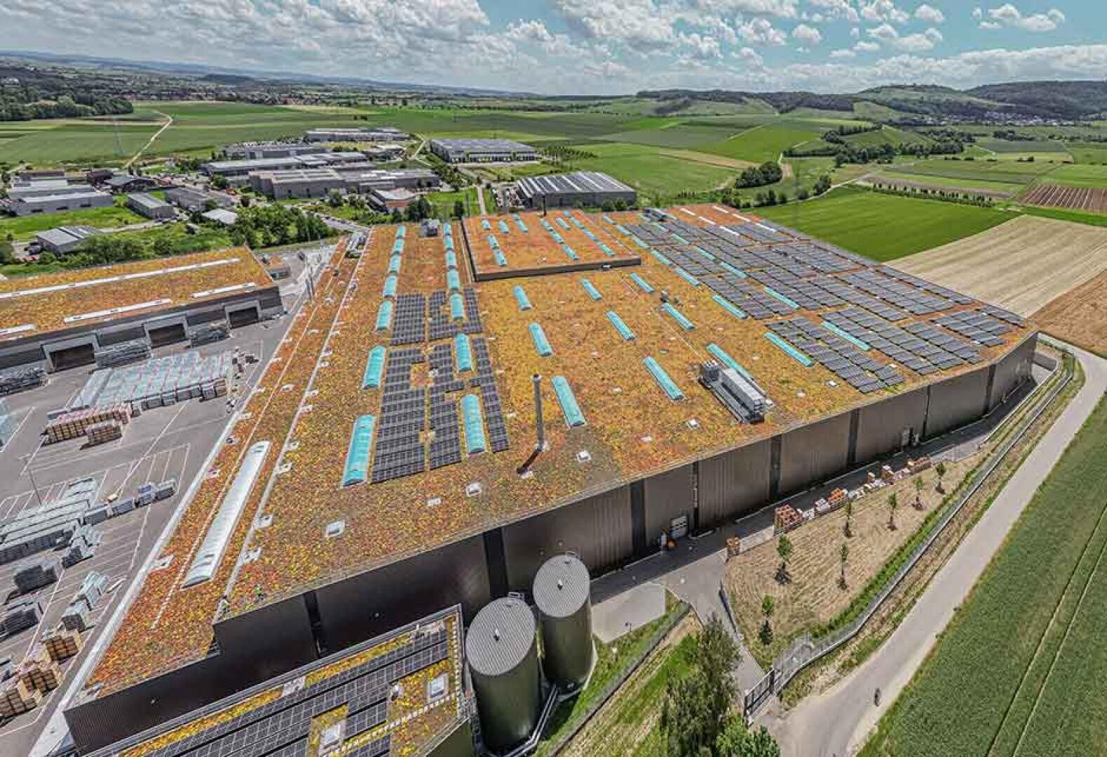 Eine große Industrielagerhalle mit einem begrünten Dach und Sonnenkollektoren, umgeben von Feldern, kleineren Gebäuden und Straßen, in einer ländlichen Landschaft unter einem teilweise bewölkten Himmel.