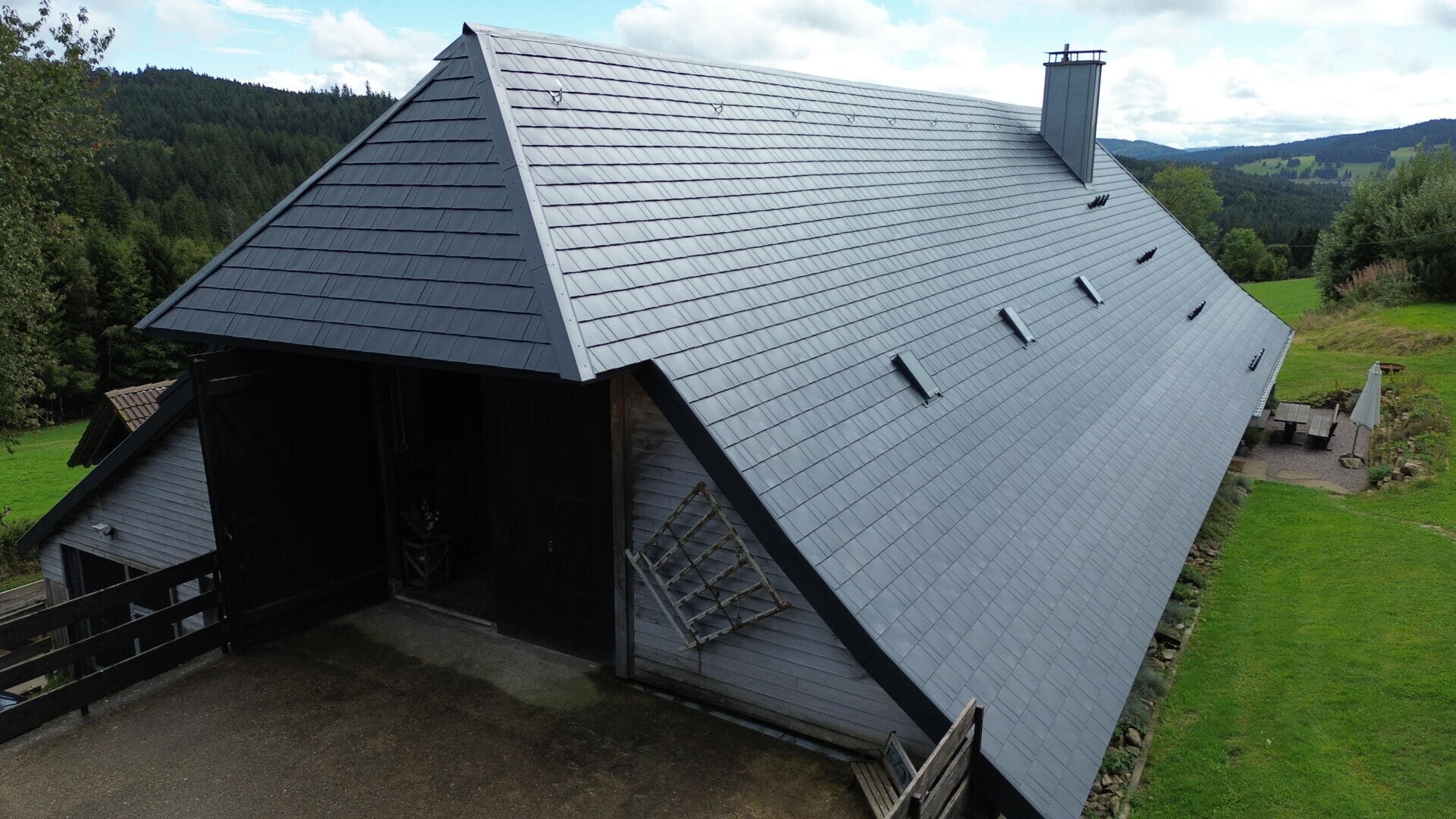 Ein großes, modernes, scheunenartiges Gebäude mit einem steilen, dunkelgrauen Schrägdach, umgeben von grünem Gras, Bäumen und Hügeln unter einem teilweise bewölkten Himmel.