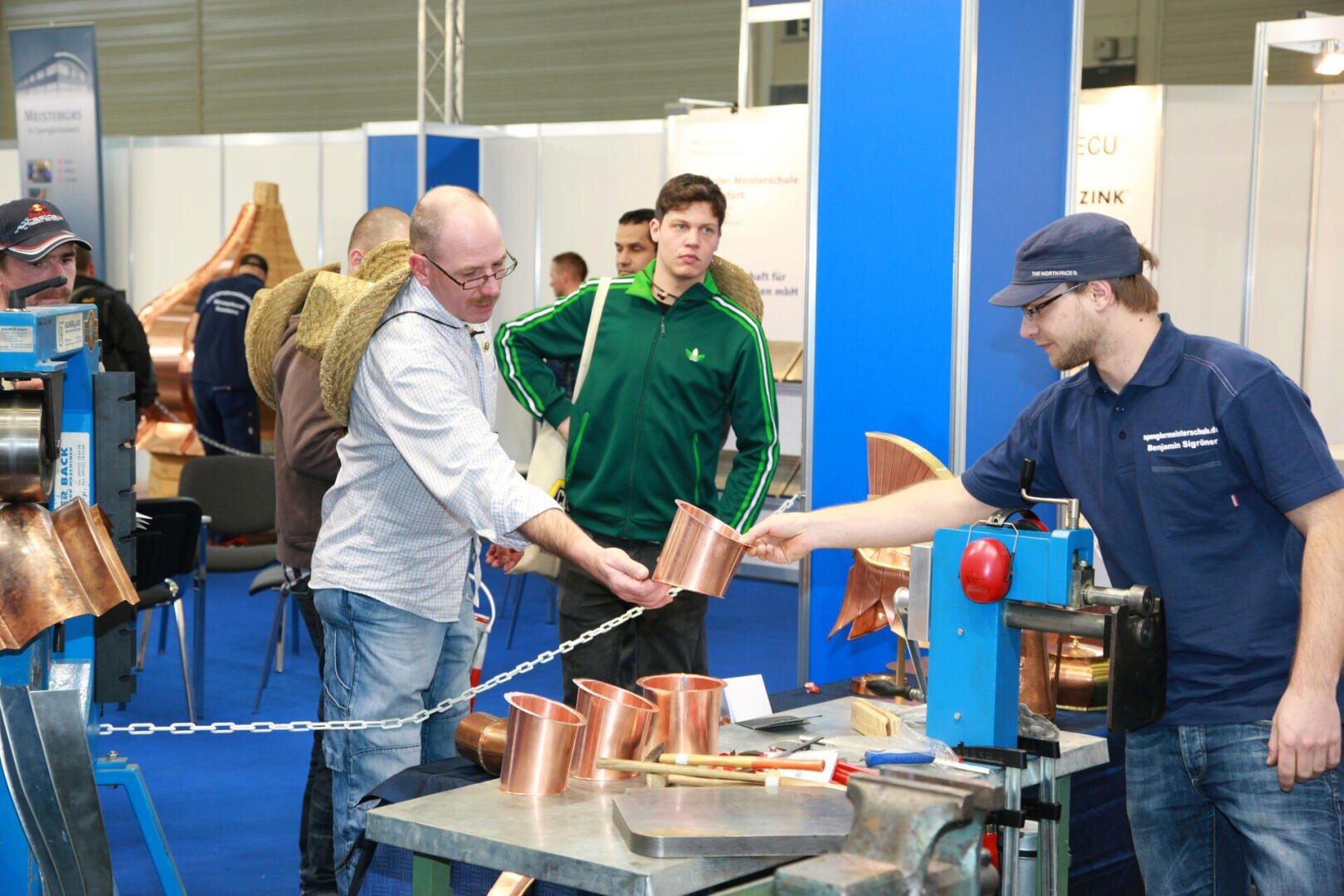 Several men stand around a display table with copper pieces at an exhibition. One man hands a copper item to another, while others observe. Blue partitions and workshop equipment are visible in the background.