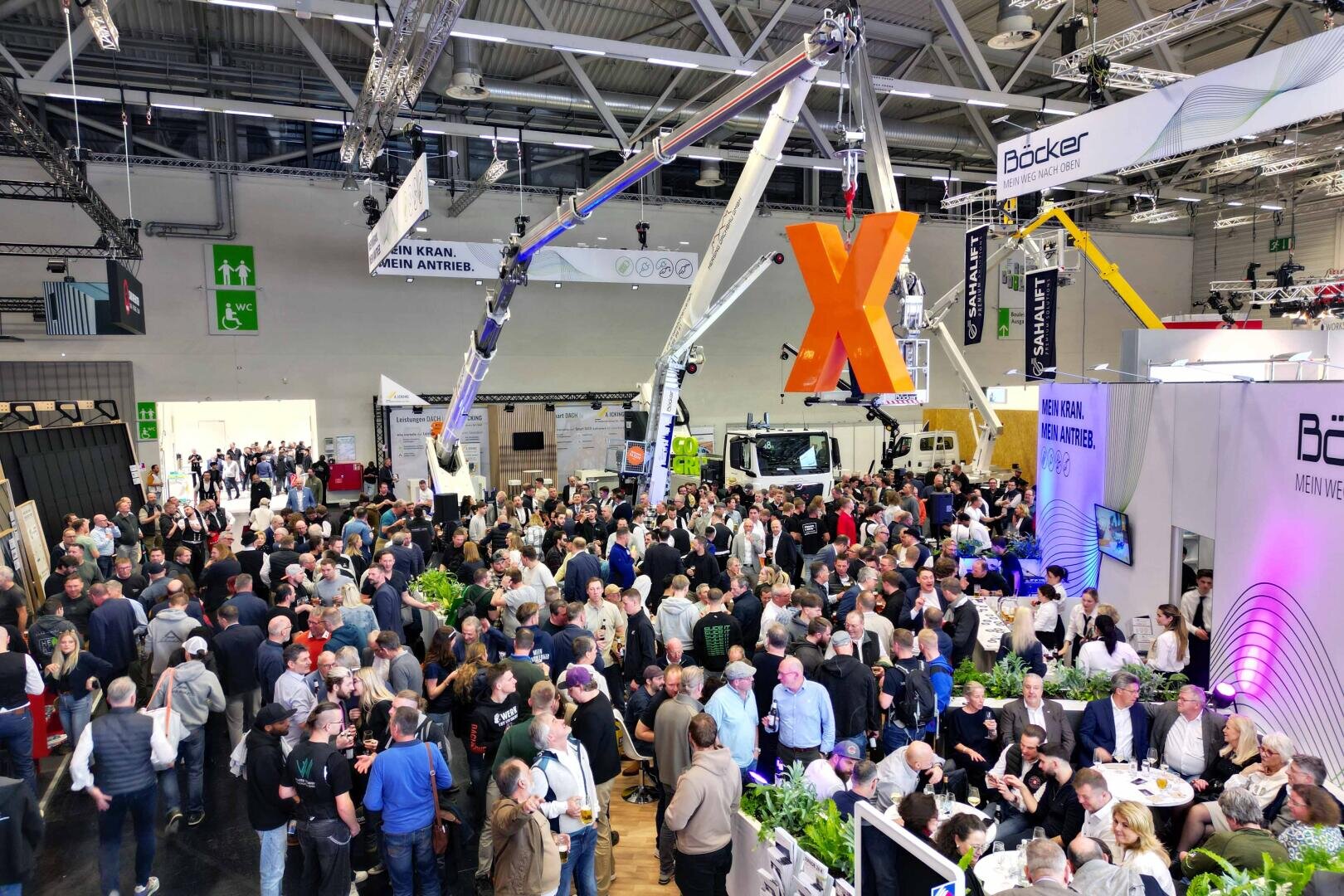 Eine große Menschenmenge besucht eine belebte Indoor-Messe mit verschiedenen Ständen, Bannern und Technologieanzeigen. Über dem Kopf hängt ein auffälliges oranges X-Schild, und im Hintergrund sind Kräne und Geräte zu sehen.