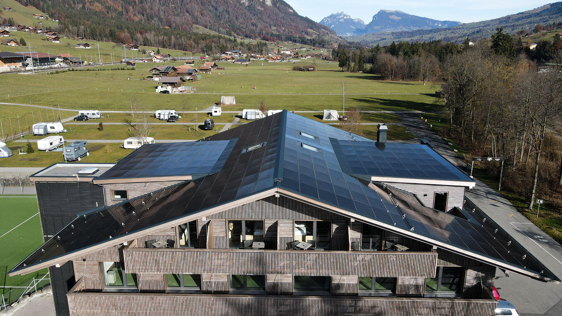 A modern building with a large, sloping roof covered with solar panels stands in a green rural area, surrounded by fields, trees, scattered houses and distant mountains under a clear sky.