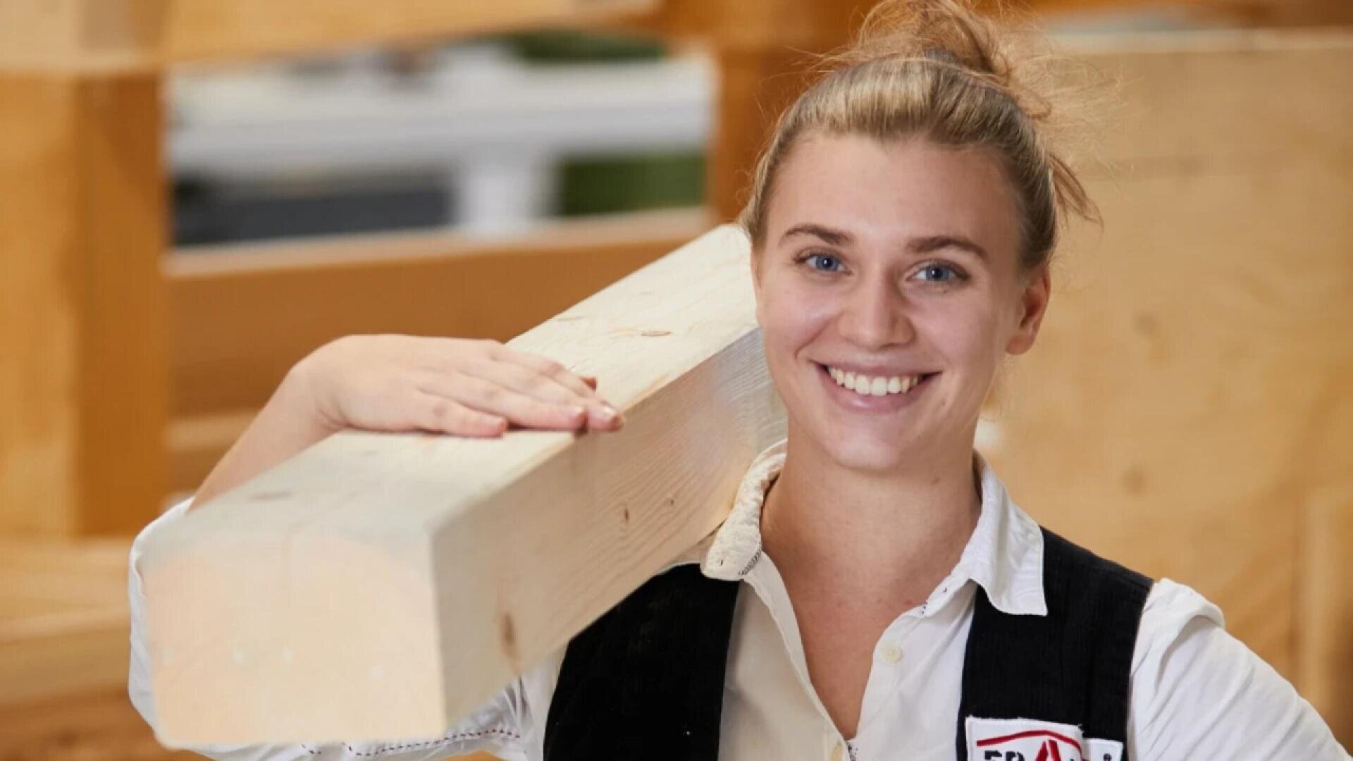 A smiling woman with blonde hair in a bun holds a large wooden beam on her shoulder. She is wearing a white shirt and a vest, standing in a carpentry workshop with wooden structures in the background.