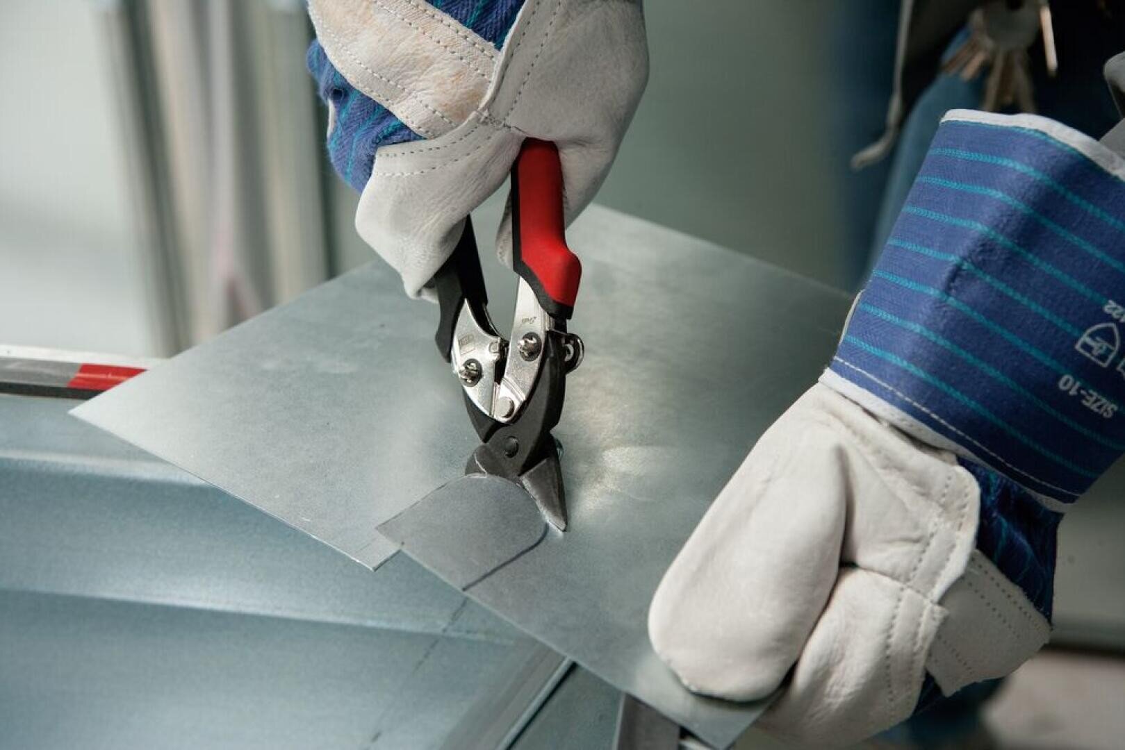 A person wearing protective gloves uses tin snips to cut a sheet of metal. The hands are focused on the cutting tool and the metal sheet, indicating a careful, precise cutting process.