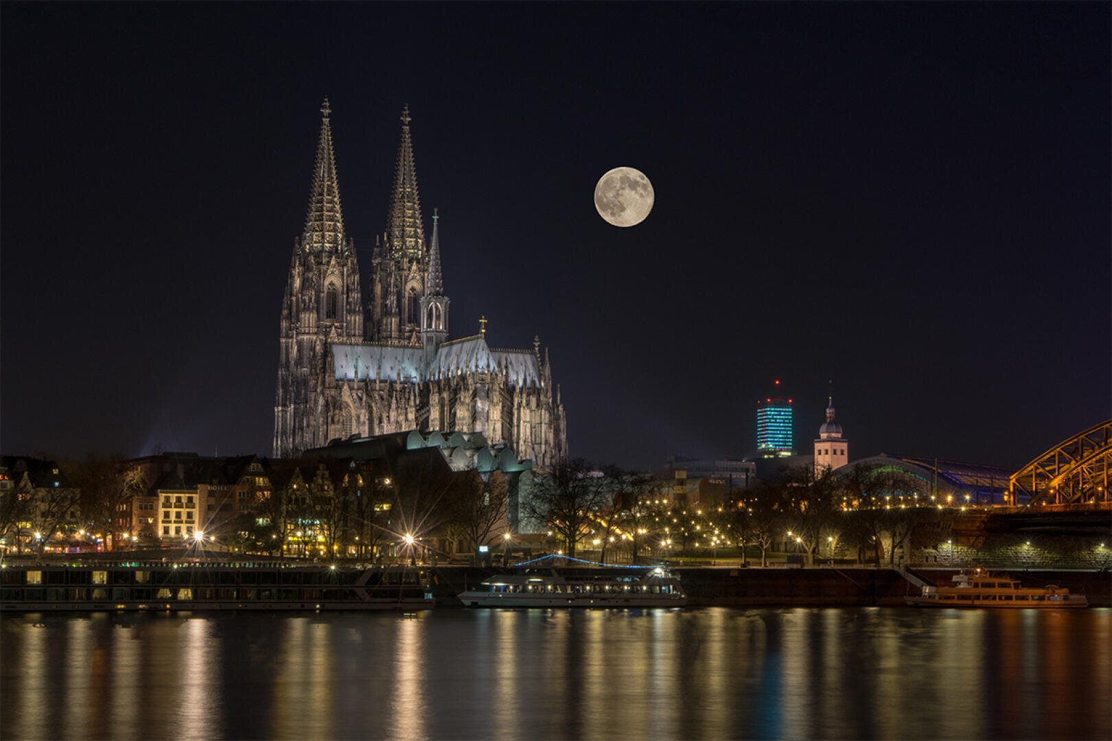 Der beleuchtete Kölner Dom steht am nächtlichen Rheinufer, über dem der Vollmond scheint. Die Lichter der Stadt spiegeln sich auf dem Wasser, und im Vordergrund liegt ein Schiff vor Anker.