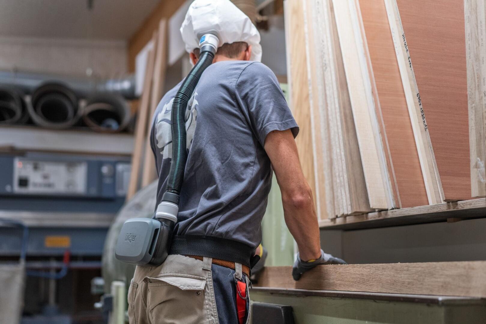 Eine Person, die eine Schutzmaske und einen am Gürtel befestigten Luftfilter trägt, prüft oder sortiert Holzbretter in einer Werkstatt oder einem Holzlager.