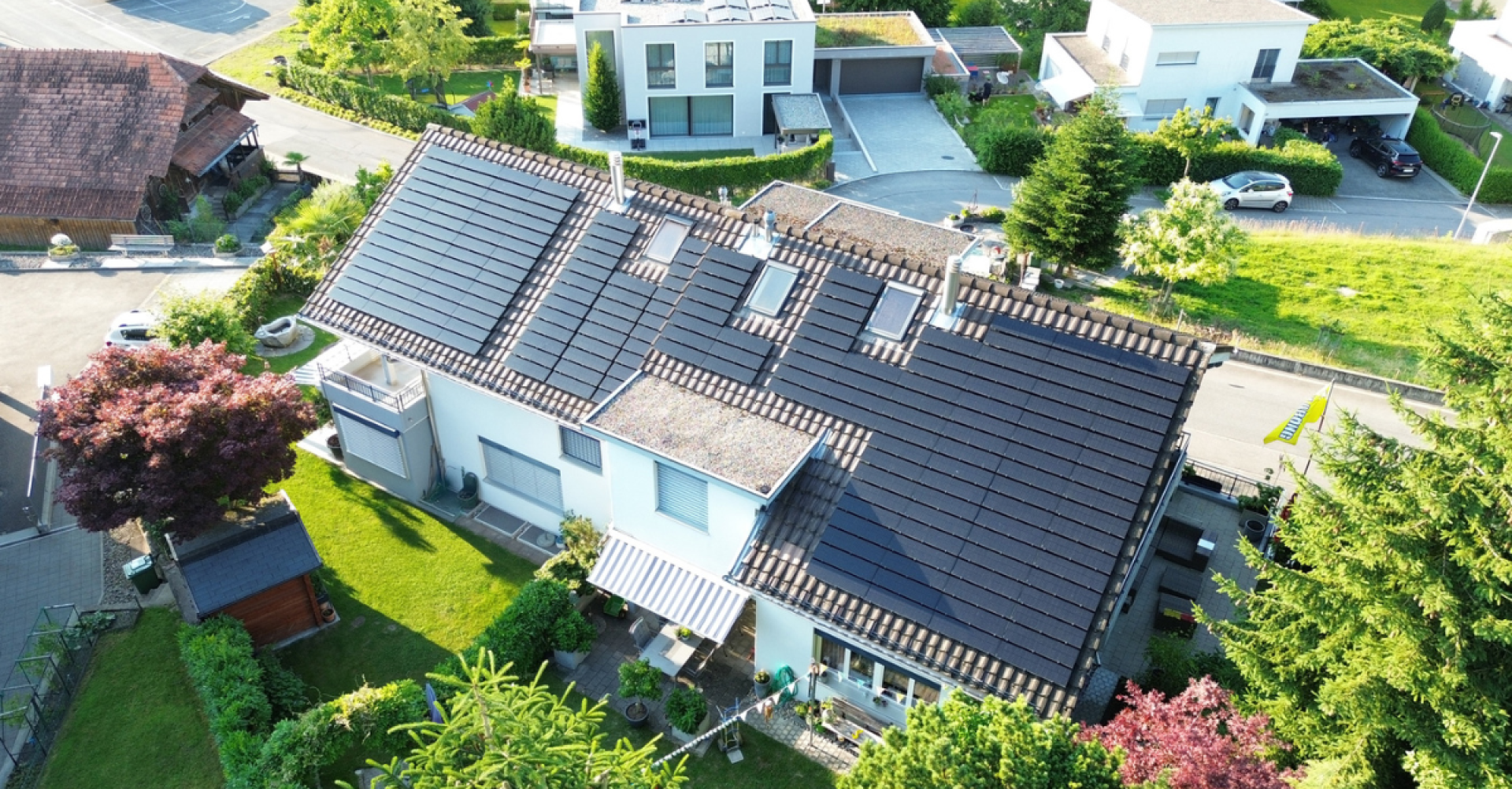 Luftaufnahme eines modernen Hauses mit einer großen Reihe von Solarzellen auf dem Dach, umgeben von einer grünen Wiese, Bäumen und benachbarten modernen Häusern in einem ruhigen Wohngebiet.