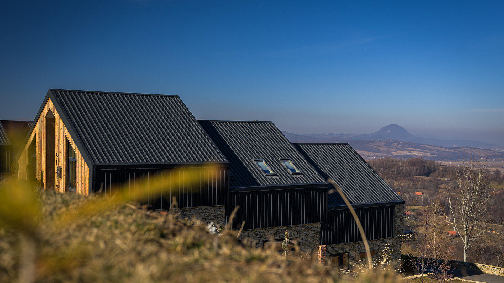 Moderne schwarze Häuser mit schrägen Metalldächern stehen auf einem grasbewachsenen Hügel unter einem strahlend blauen Himmel, mit Bergen und Tälern in der Ferne im Hintergrund.