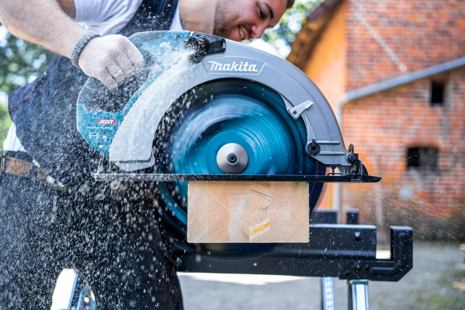 Eine Person schneidet mit einer Makita-Kreissäge ein Holzbrett im Freien; Holzstaub und Sägespäne werden in die Luft gesprüht, wenn sich das Blatt schnell dreht.