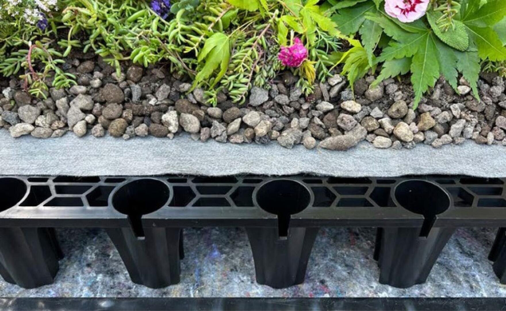 Cross-section of a green roof system with soil, plants, gravel, a fabric layer and a drainage layer made of black plastic with vertical supports.