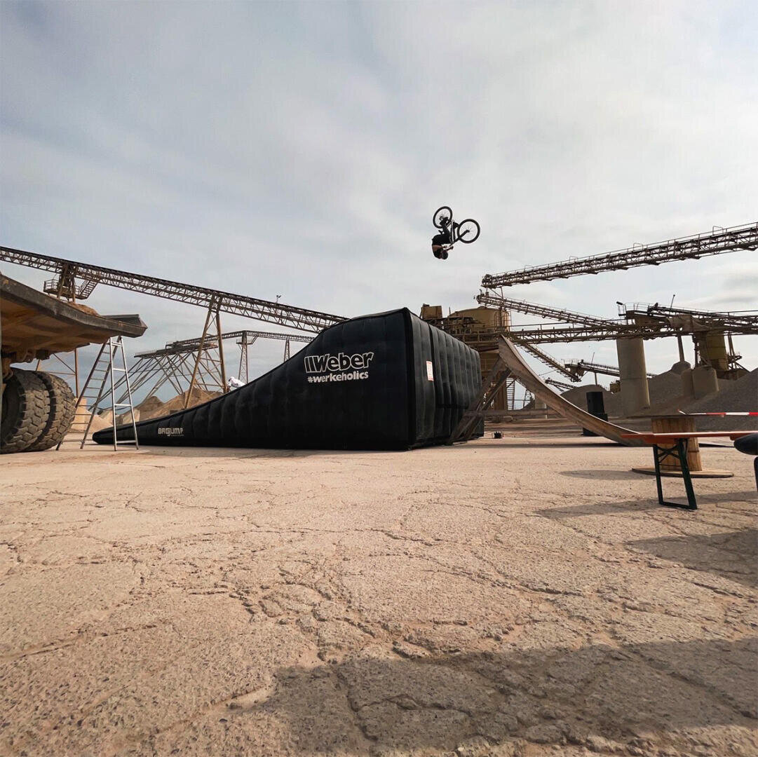 A person on a bicycle performs a trick in the air above an inflatable runway in an industrial environment with conveyor belts and machines in the background.