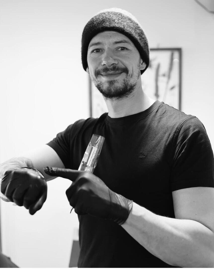 A man in a black cap, black T-shirt and black gloves smiles while holding a tattoo machine and standing in a room with framed artwork in the blurred background.