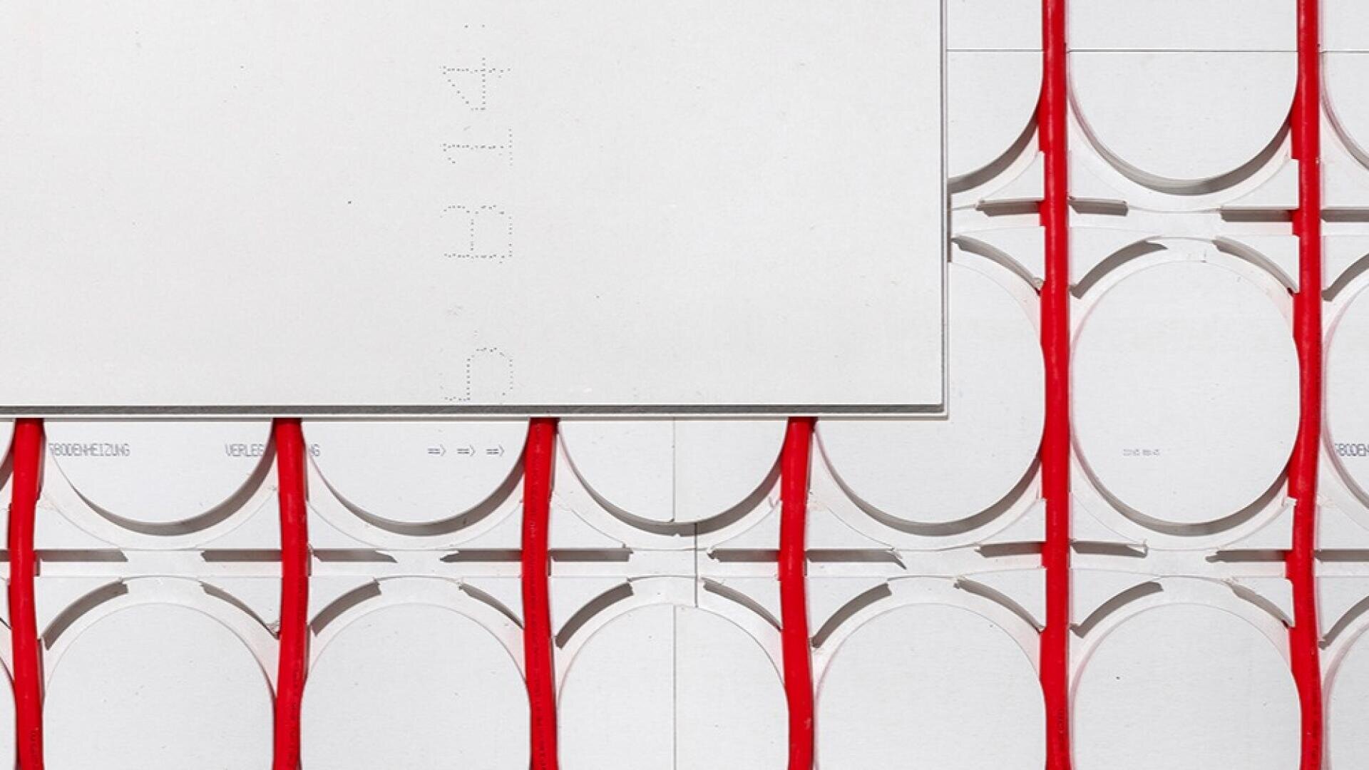 Ein weißes Paneel ist über einem Raster aus kreisförmigen Ausschnitten und roten vertikalen Rohren angebracht und verdeckt teilweise das geometrische Muster an der Wand dahinter.