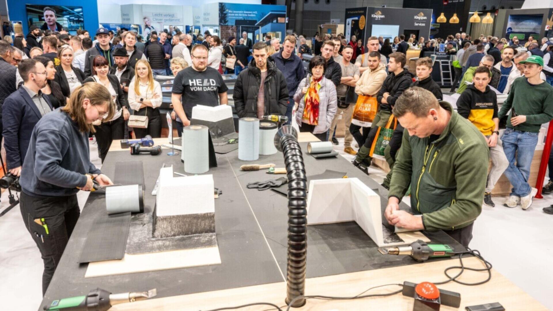 A crowd watches two people demonstrate roofing or construction techniques at a trade show booth, using tools and construction materials on a table. Various supplies and equipment are visible, with many onlookers observing closely.