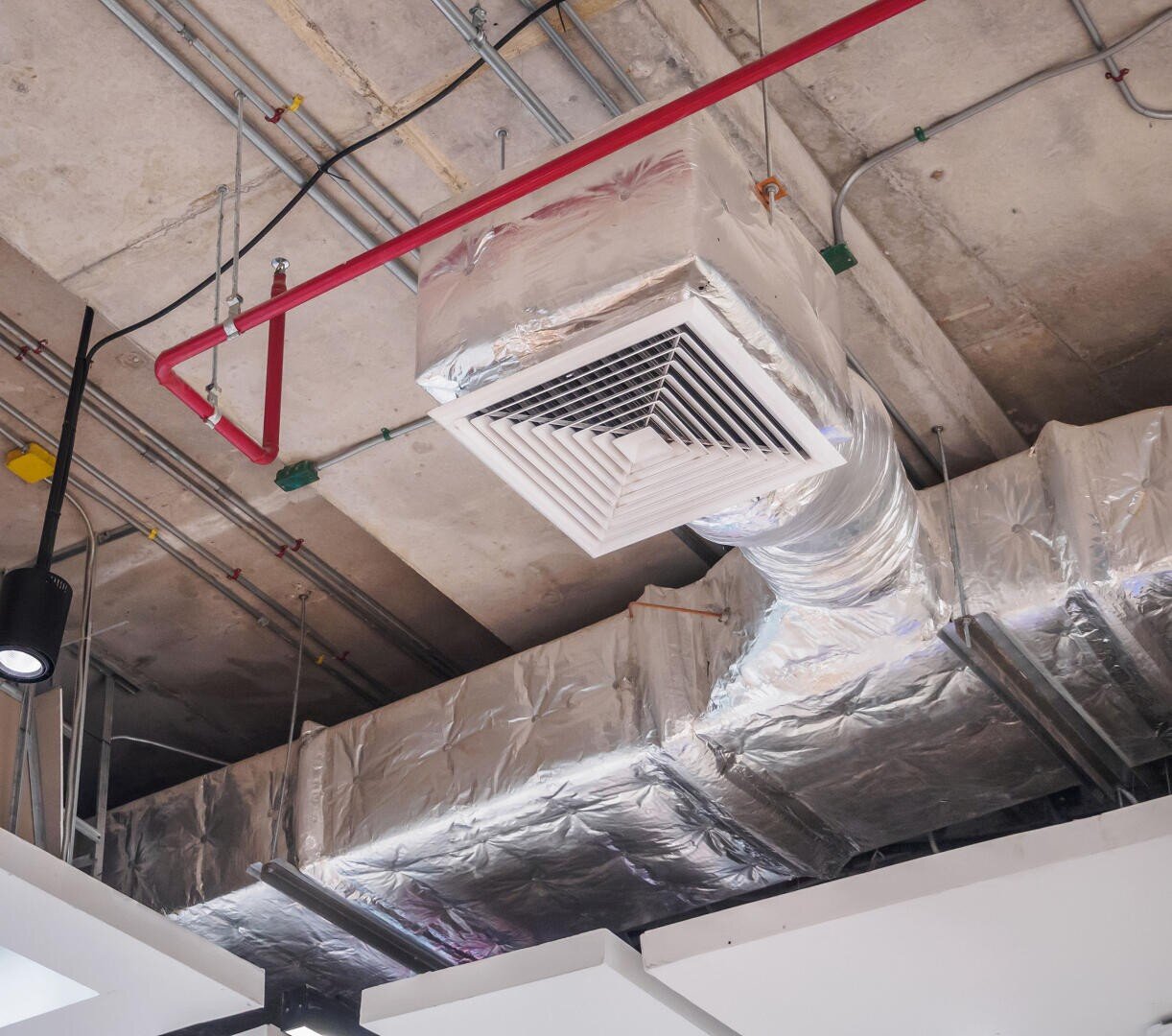 Exposed air duct system with a vent, wrapped in reflective insulation, on an unfinished concrete ceiling with visible pipes, electrical conduits, and light fixtures.