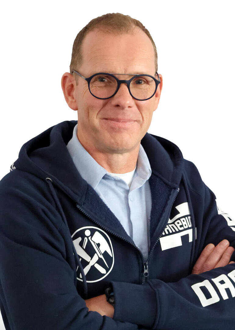 Ein Mann mit kurzen hellen Haaren und Brille, der einen blauen Kapuzenpullover mit weißer Aufschrift über einem hellblauen Kragenhemd trägt, steht mit verschränkten Armen und lächelt leicht vor einem weißen Hintergrund.