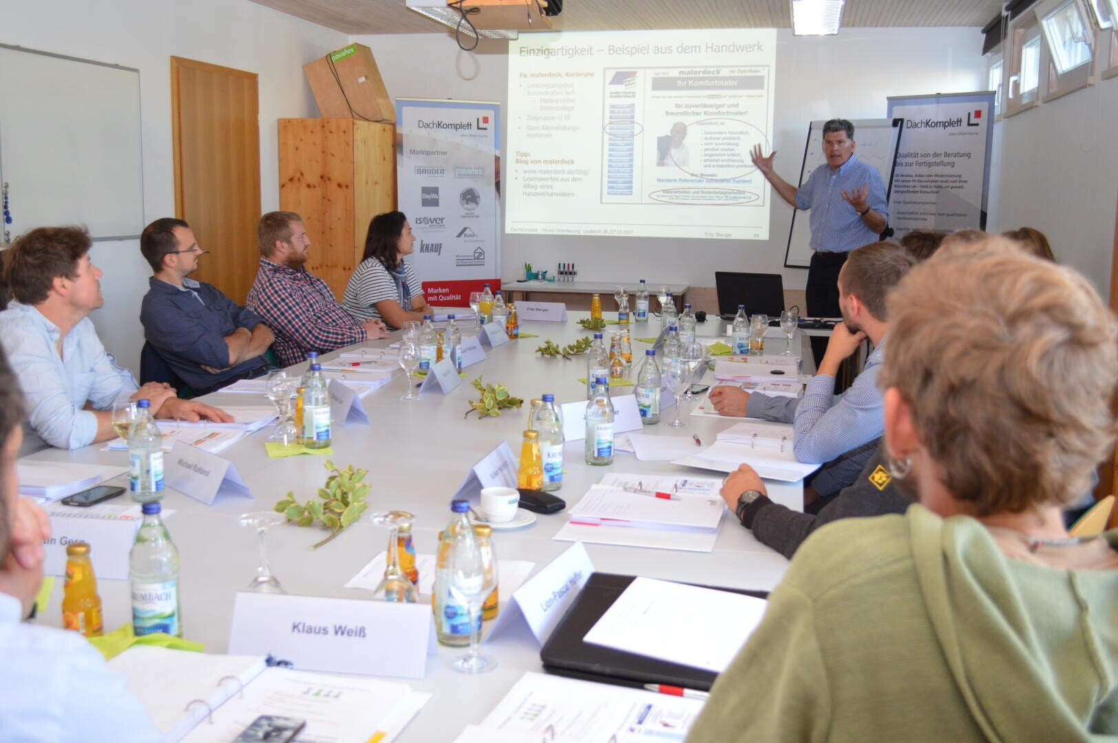 Eine Gruppe von Personen sitzt mit Papieren und Getränken um einen Konferenztisch und hört einem Moderator zu, der in einem hellen Besprechungsraum vor einem Bildschirm steht, auf dem eine Präsentation gezeigt wird.