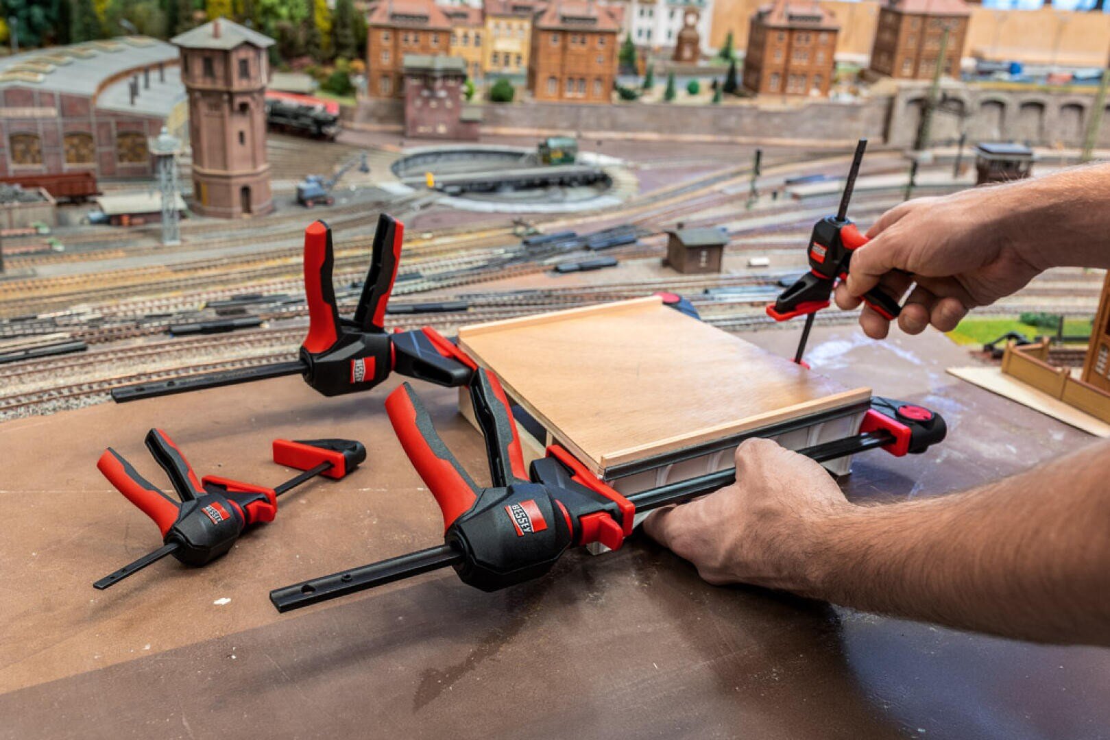 A person uses clamps to secure wooden pieces for a model train layout while tightening screws, with tracks, trains, and buildings visible in the detailed background.