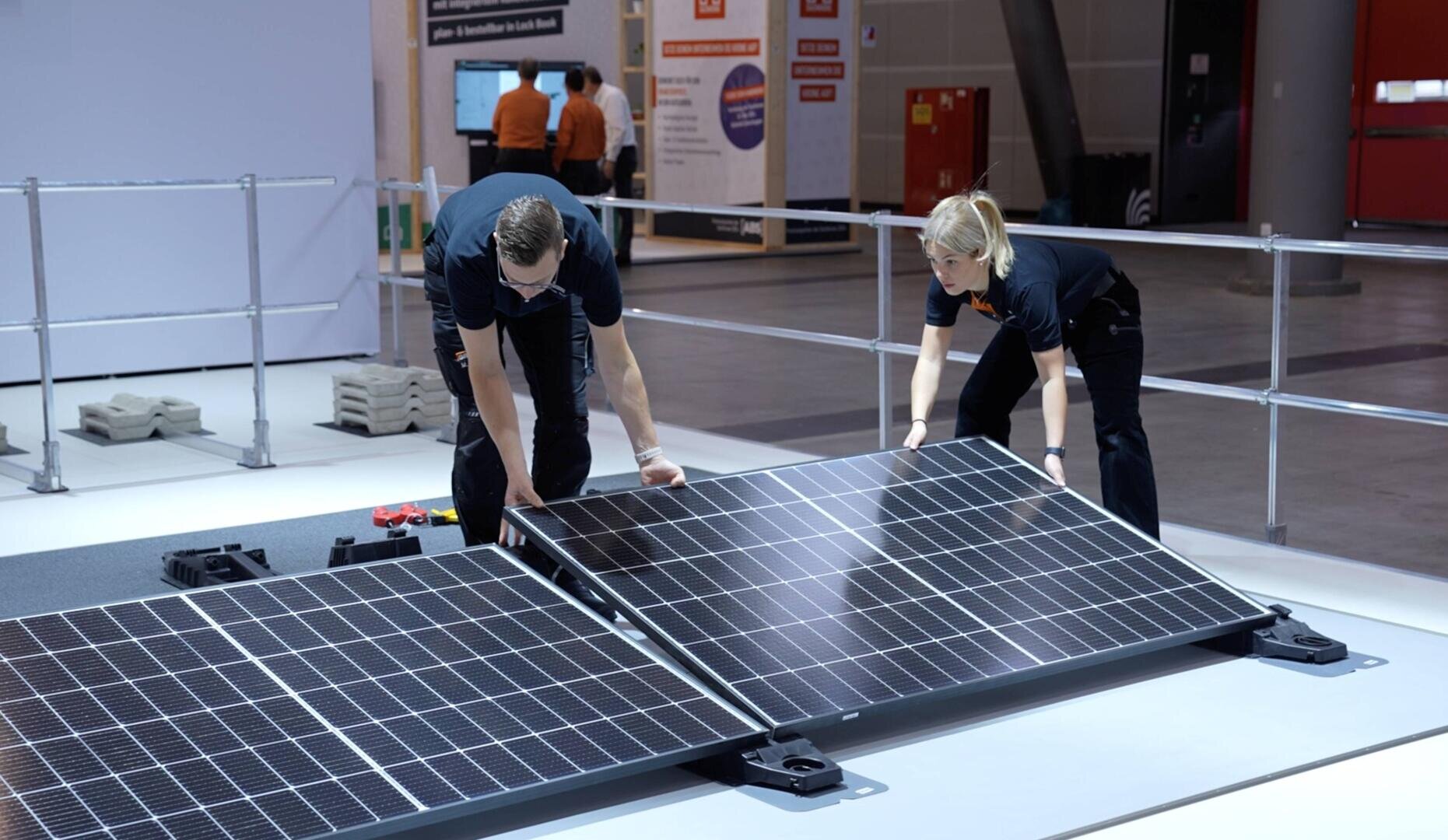 Two people in black shirts are installing large solar panels indoors on a white floor, with exhibition booths and more people visible in the background.