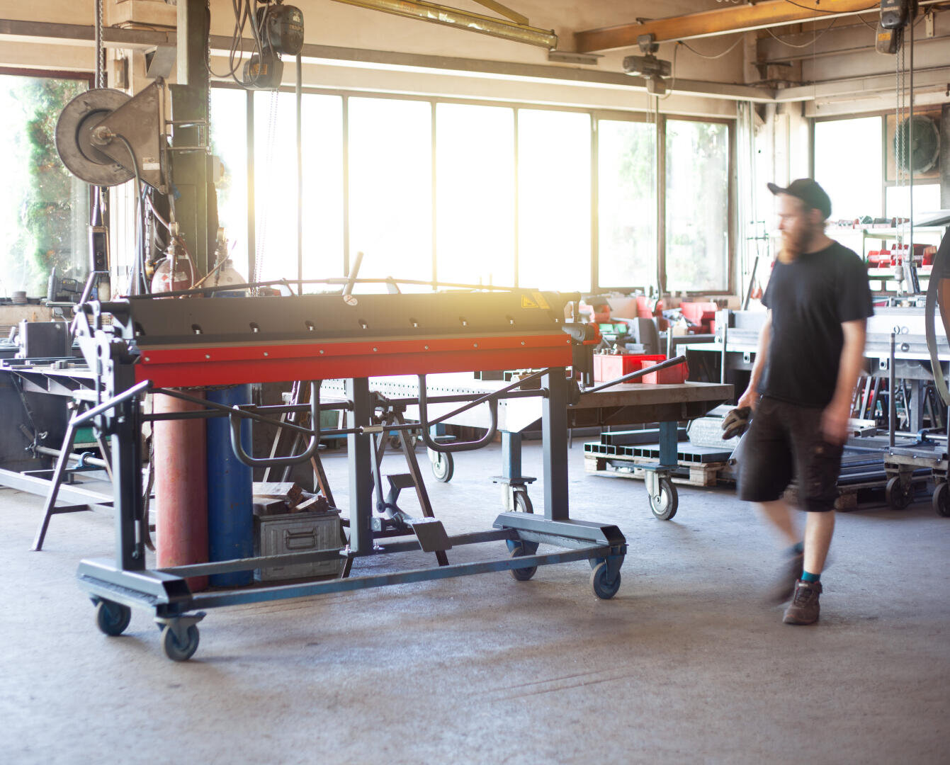 Ein Mann mit einer Mütze und dunkler Kleidung geht durch eine helle Werkstatt voller Geräte, darunter eine große Metallmaschine auf Rädern in der Mitte und verschiedene Werkzeuge auf Bänken an den Fenstern.