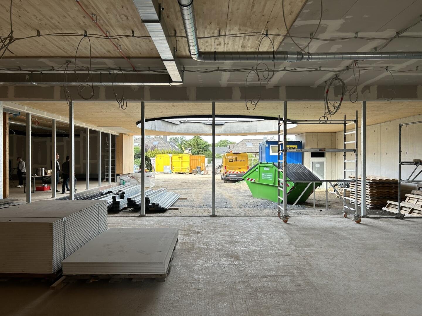 Blick aus dem Inneren eines im Bau befindlichen Gebäudes mit freiliegenden Drähten, gestapelten Materialien und einem offenen Bereich, der zu einem Außenbereich mit Containern, Baumaschinen und einigen Menschen im Hintergrund führt.