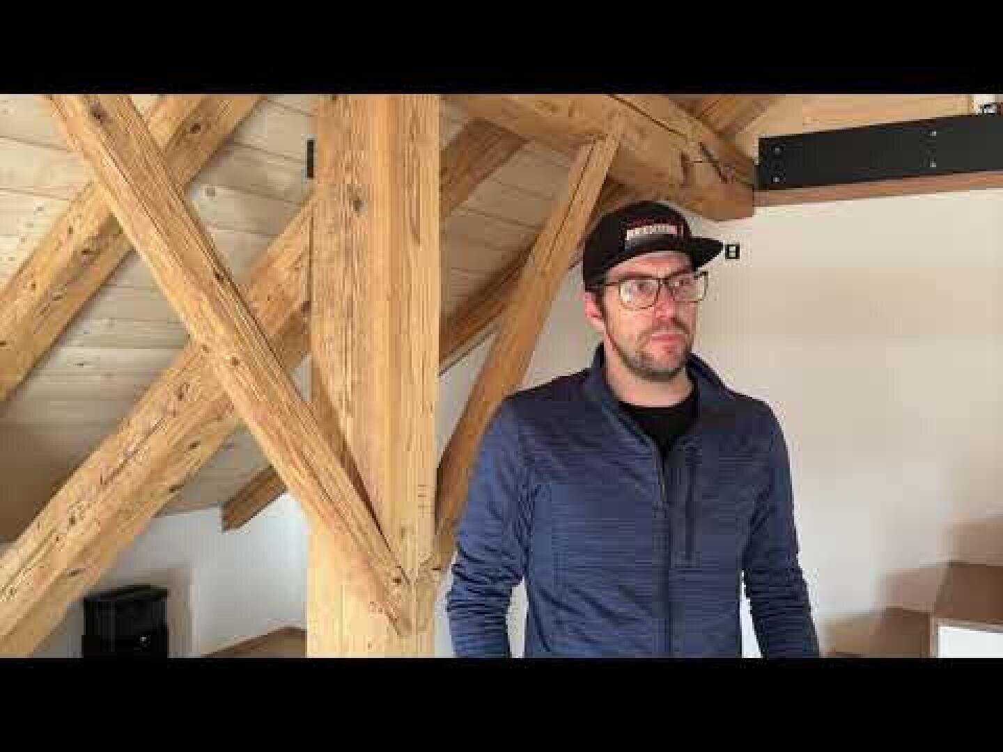 Ein Mann, der eine Brille, eine dunkle Baseballkappe und eine blaue Jacke mit Reißverschluss trägt, steht in einem Raum mit freiliegenden Holzbalken und einer schrägen Decke. Der Raum scheint ein Dachboden oder ein Loft mit hölzernen Elementen zu sein.