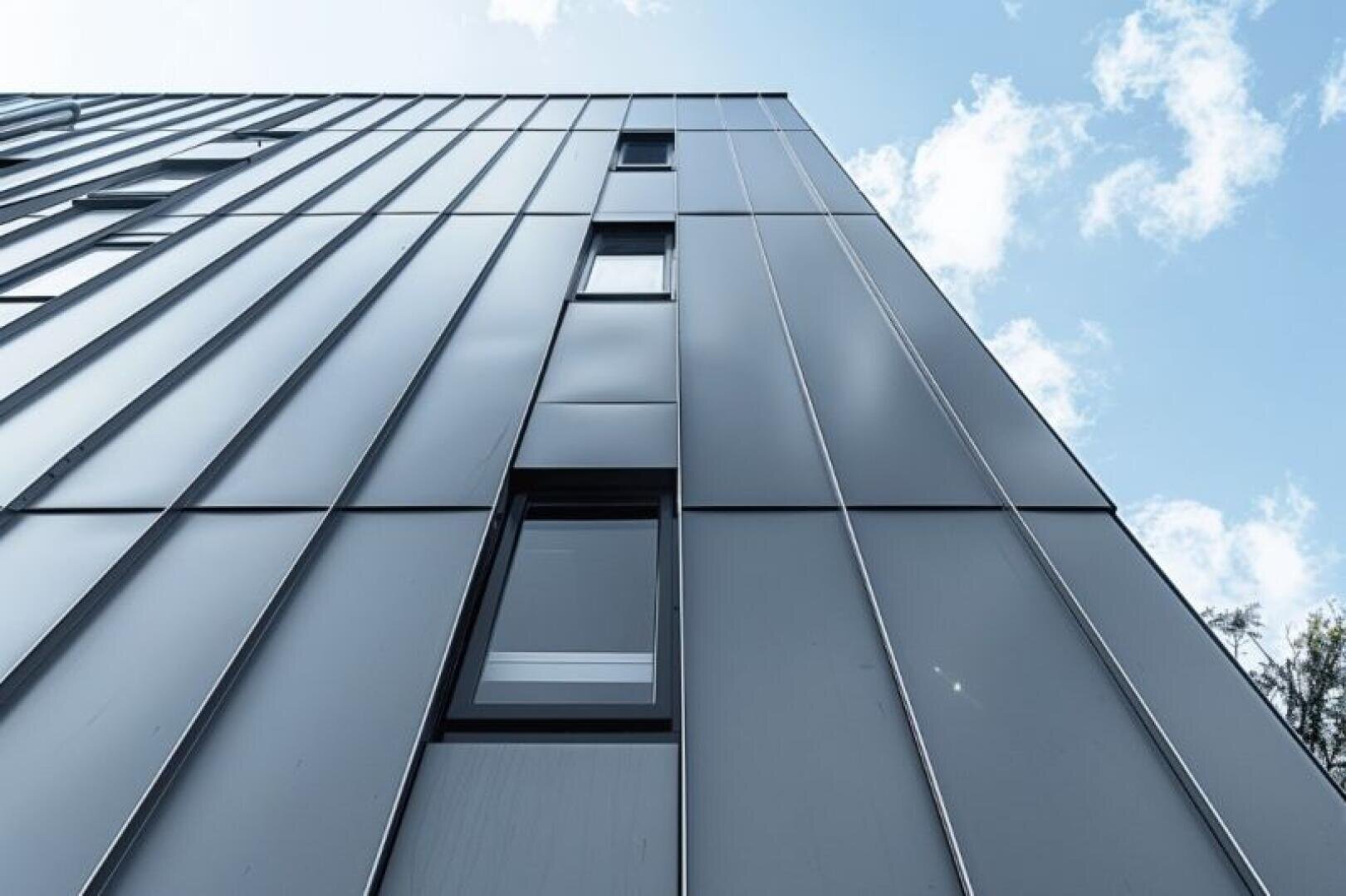 Blick aus geringer Höhe auf ein modernes Gebäude mit glatten Metallpaneelen und mehreren schmalen Fenstern vor einem blauen Himmel mit vereinzelten Wolken.