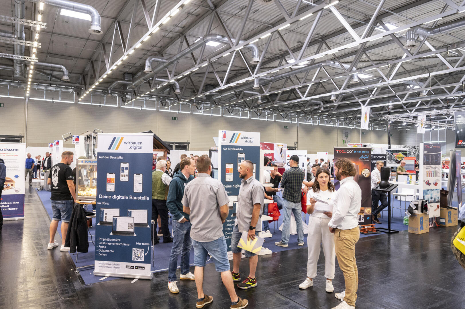 At a busy indoor trade fair or exhibition, people exchange ideas and browse the stands. Various displays, banners and information stands can be seen under a large industrial ceiling with bright lights.