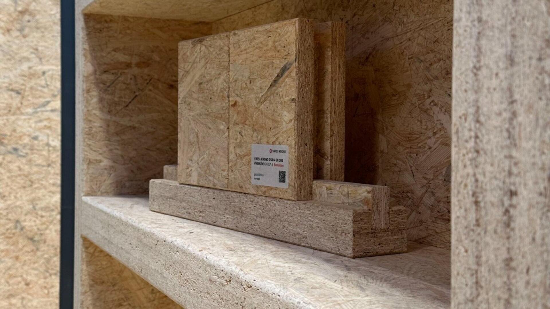 A close-up of a shelf holding samples of oriented strand board (OSB) and particleboard, with a small label attached to one sample. The background and shelf are also made of similar wood panels.
