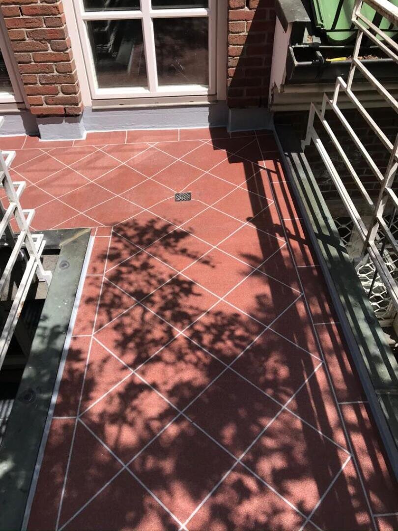 Sonnenlicht und Baumzweige werfen Schatten auf einen rot gefliesten Balkon mit weißen Fugen, der von einem weißen Geländer begrenzt wird. Im Hintergrund sind ein Fenster und eine Backsteinmauer zu sehen.