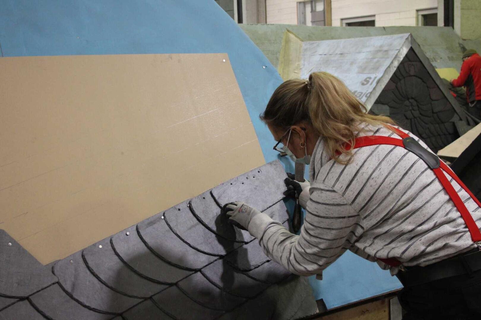 Eine Person, die Handschuhe, Schutzbrille und ein gestreiftes Hemd trägt, verlegt dunkle Schindeln auf einem Schrägdachmodell in einem Innenraum und konzentriert sich sorgfältig auf ihre Arbeit.