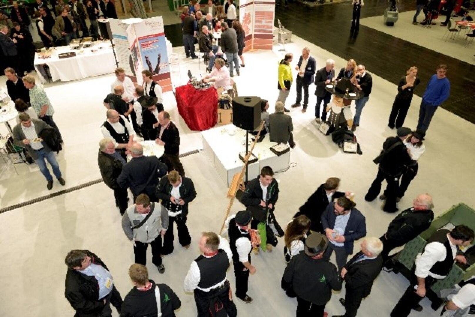 A crowded event in a building with people standing in groups and chatting. Tables with food and drinks can be seen, and attendees are wearing business and casual attire. Some people appear to be socializing or chatting.