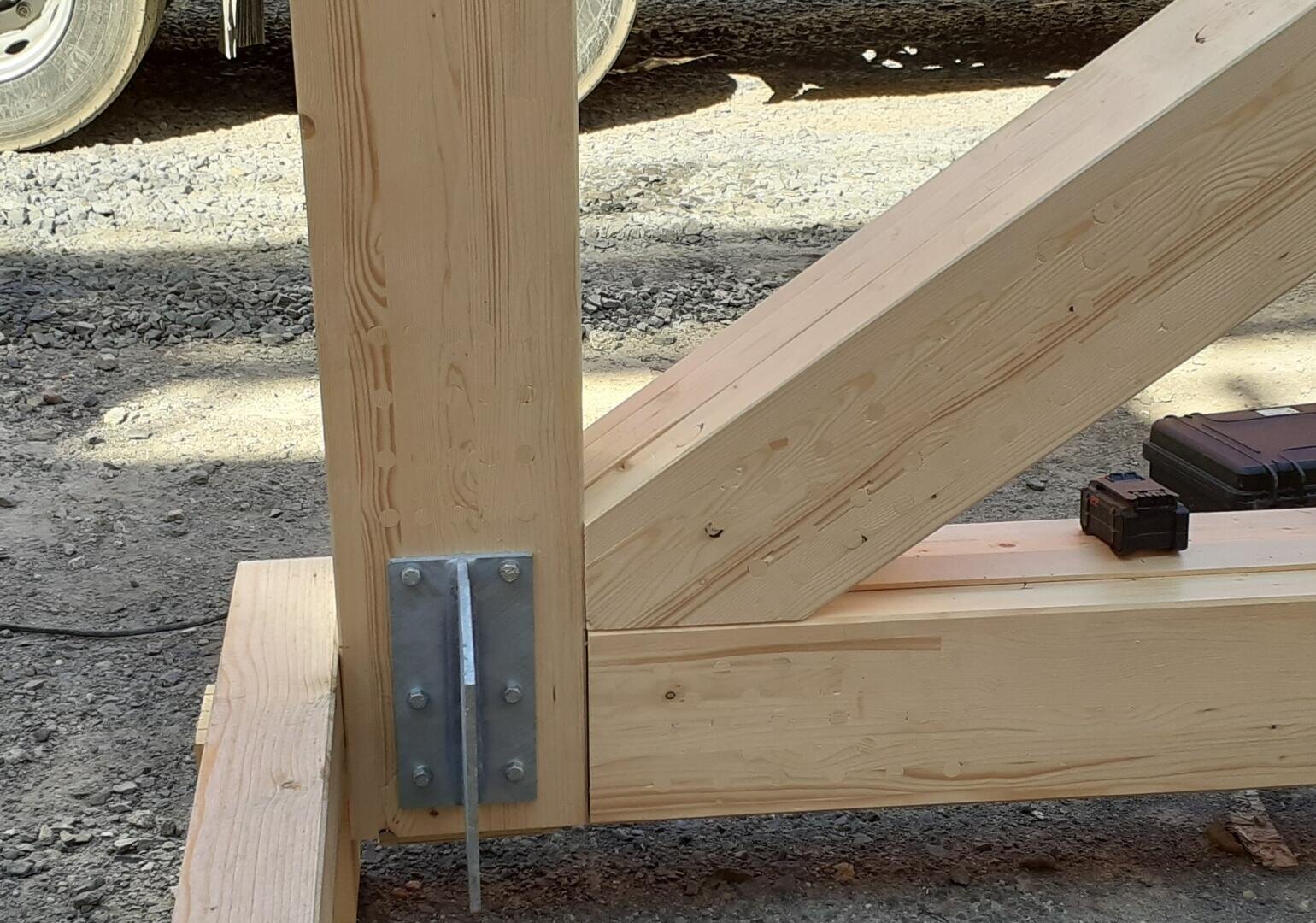 Nahaufnahme einer Holzbalkenkonstruktion, die mit Metallplatten und Bolzen zusammengefügt ist und auf einer Kiesfläche ruht. Im Hintergrund sind teilweise ein Elektrowerkzeug und ein Rad zu sehen.