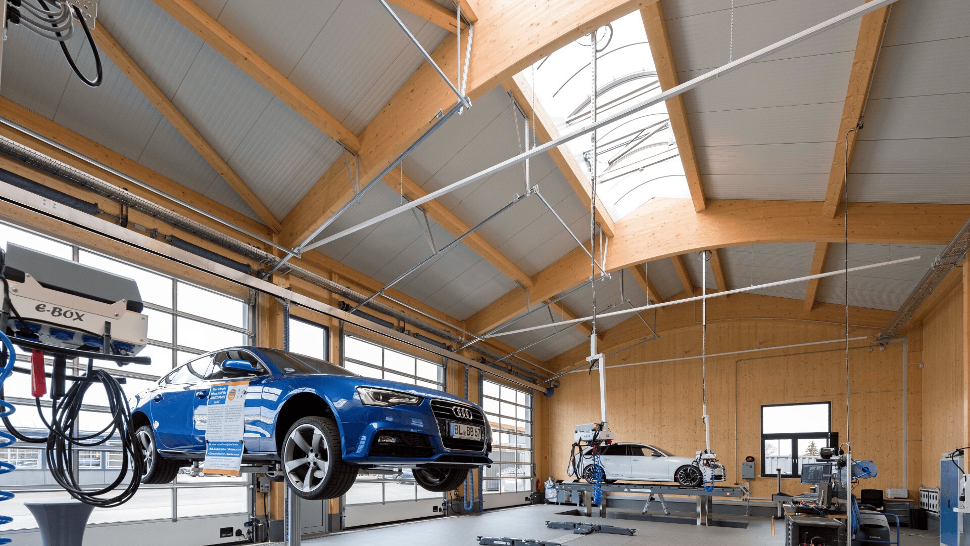 Eine moderne, geräumige Autowerkstatt mit hohen Holzdecken, großen Fenstern und zwei Autos – einem blauen Audi auf einer Hebebühne und einem weiteren weißen Auto im Hintergrund. Geräte und Werkzeuge sind überall sichtbar.