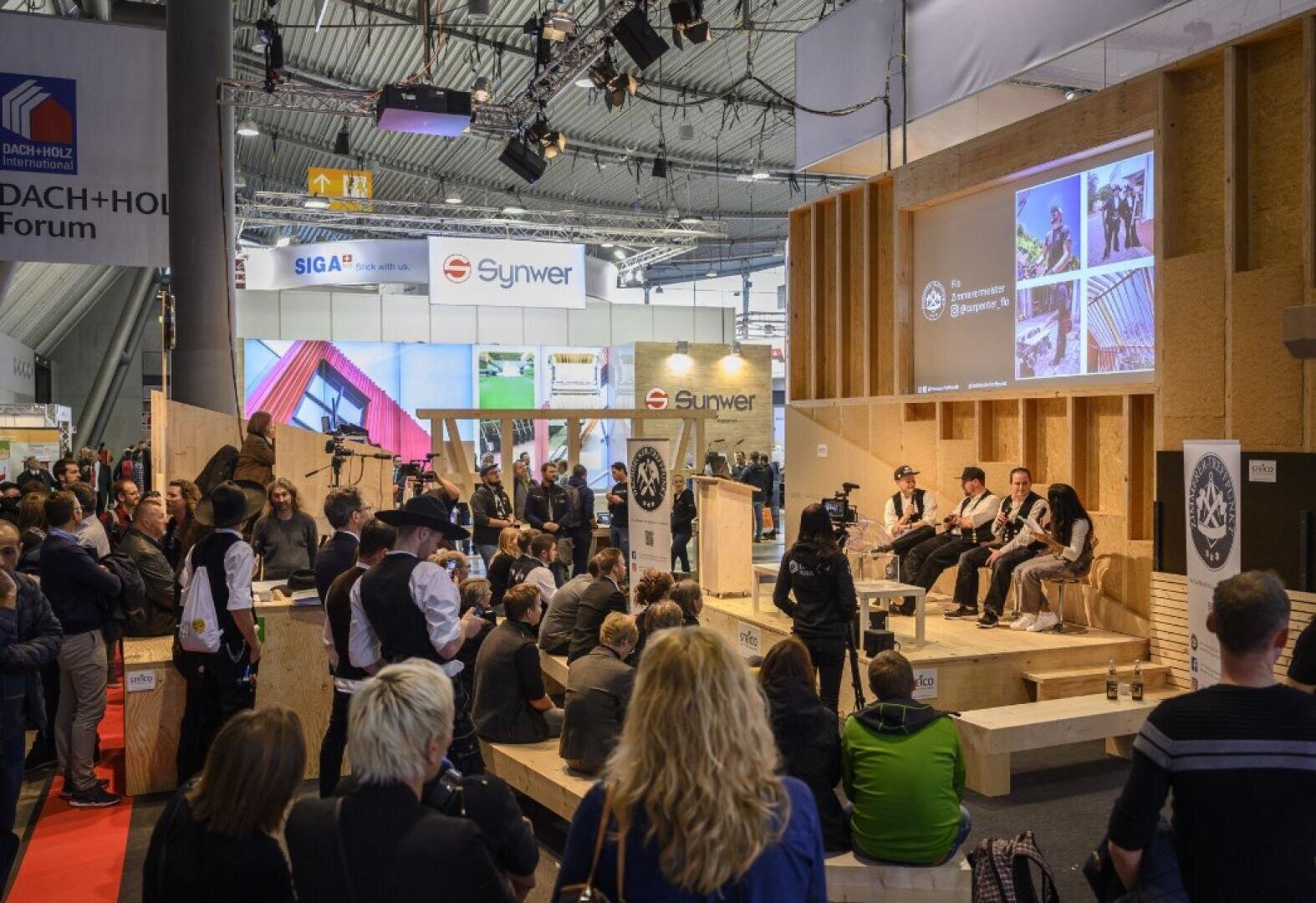 Eine Menschenmenge verfolgt eine Podiumsdiskussion bei einer Veranstaltung der Baubranche in einer großen Ausstellungshalle. Die Redner sitzen auf einer Holzbühne und im Hintergrund sind Firmenstände zu sehen.