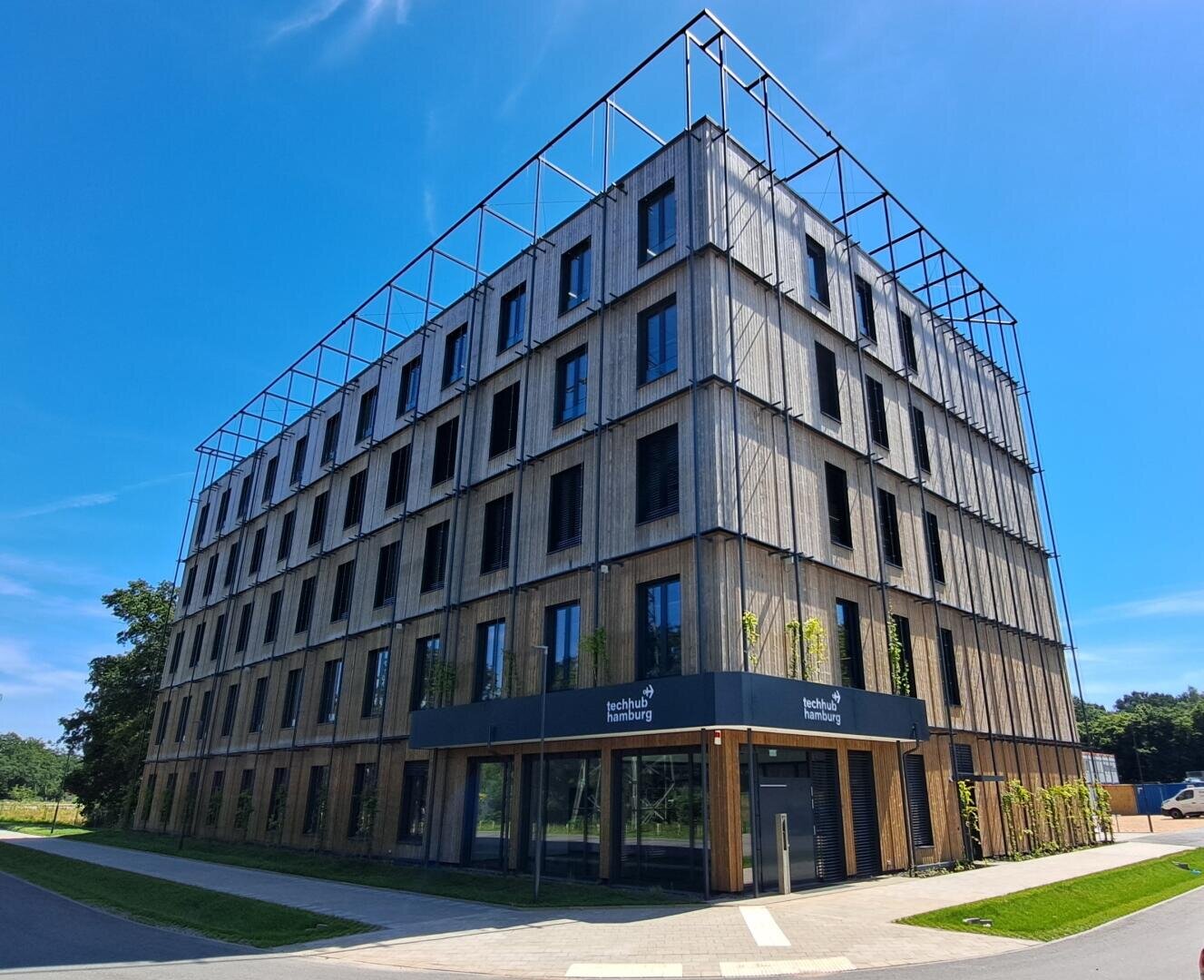 Ein modernes, vierstöckiges Bürogebäude mit großen Fenstern und einer holzgetäfelten Fassade, auf der das Logo der Technischen Hochschule Ingolstadt vor einem strahlend blauen Himmel zu sehen ist.