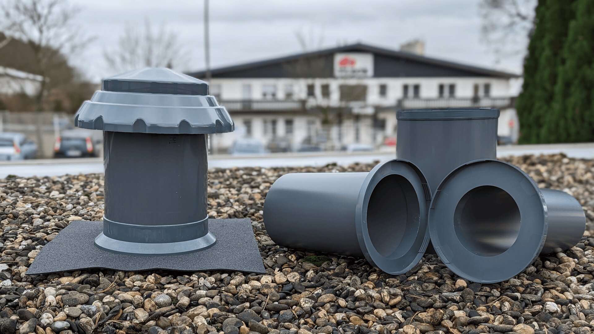 Auf einem Kiesdach sind Dachentlüftungs- und Rohrverbindungen aus grauem Kunststoff zu sehen, im Hintergrund sind unter einem bewölkten Himmel ein verschwommenes Gebäude und Bäume zu sehen.