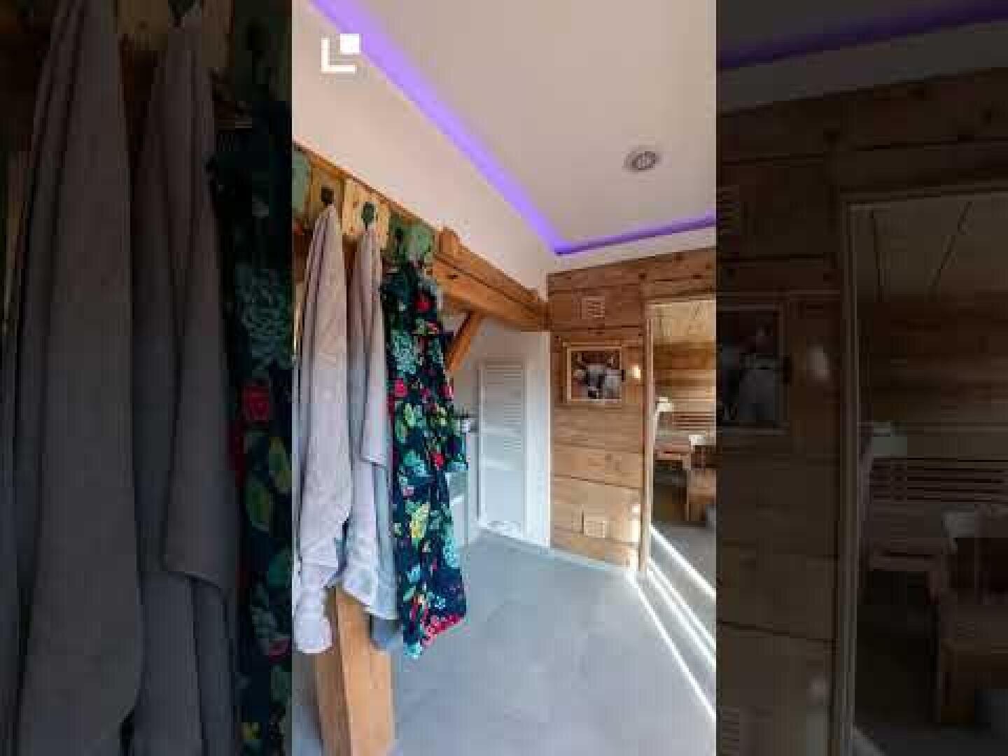 Ein gemütliches Holzzimmer mit Bademänteln und Handtüchern, die an Haken hängen, hellem Tageslicht, LED-Leuchtbändern an der Decke und einem Blick in ein anderes rustikales Zimmer durch eine offene Tür.