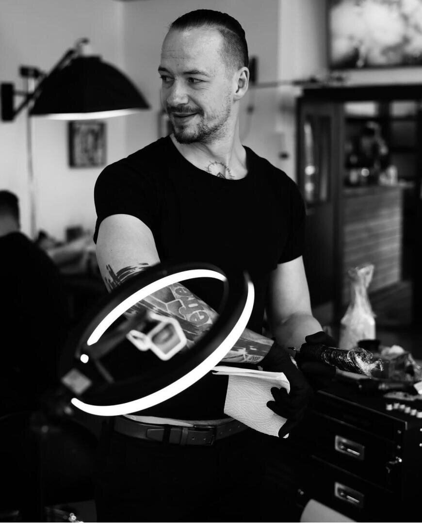 A tattoo artist with a tattooed arm stands in a studio, holding a paper towel and wearing gloves. A ring light illuminates his arm and he looks to the side with a smile. The picture is in black and white.