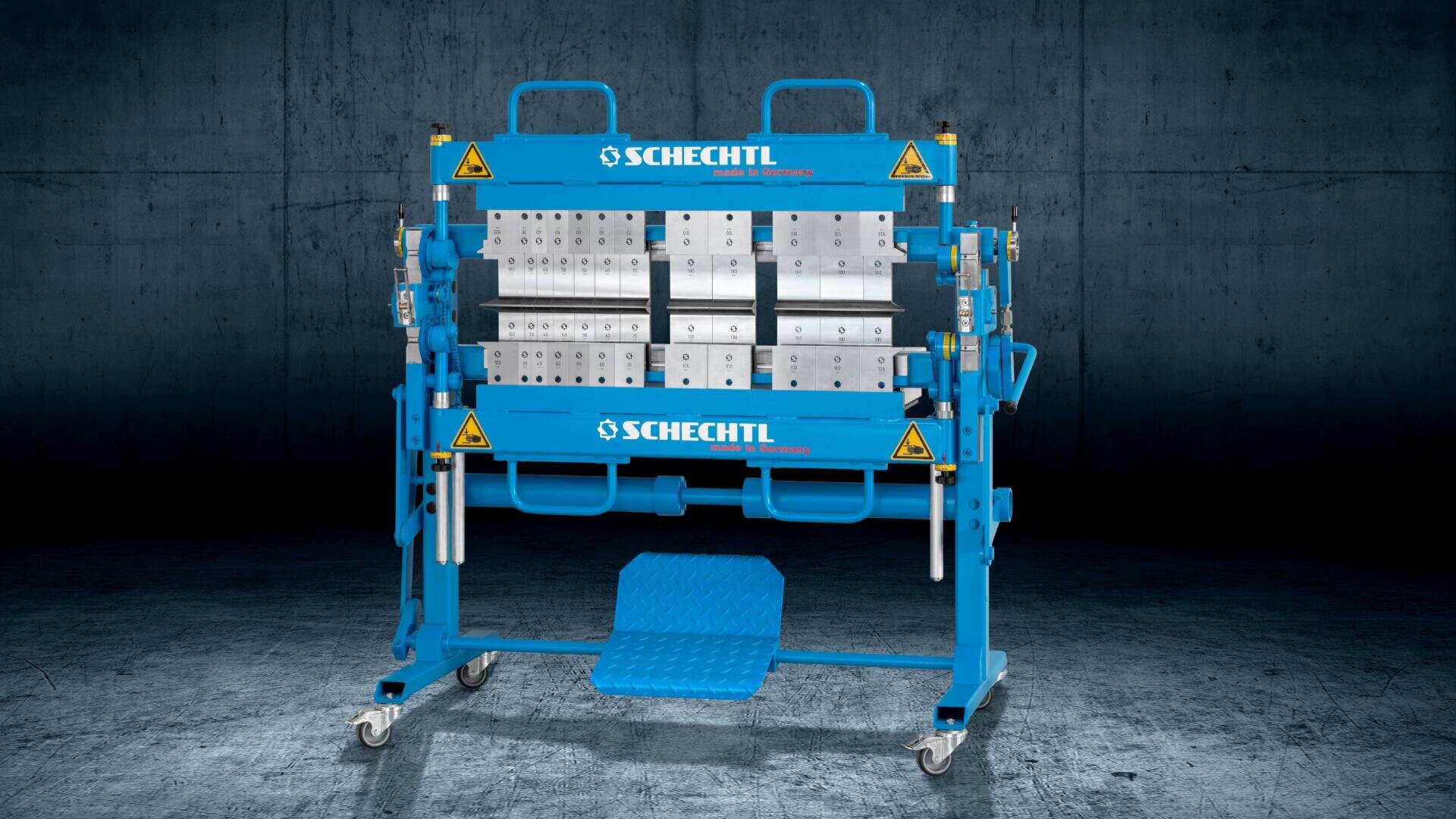 Eine blaue Schechtl-Metallbiegemaschine mit verstellbaren Komponenten und Warnschildern steht auf Rädern vor einem dunklen industriellen Hintergrund.